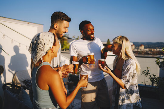 Young diverse adults laughing and socializing with beer mugs at a golden hour rooftop party