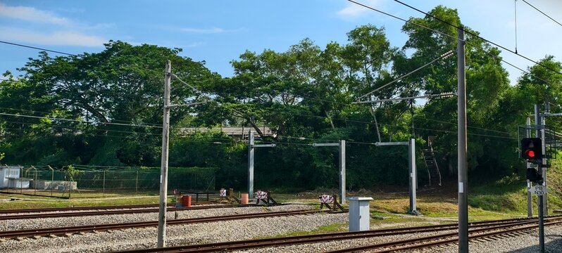 Railway yard infrastructure with multiple parallel train tracks, buffer stops, and overhead electrical wires supported by metal poles. A red railway signal light is visible on the right.