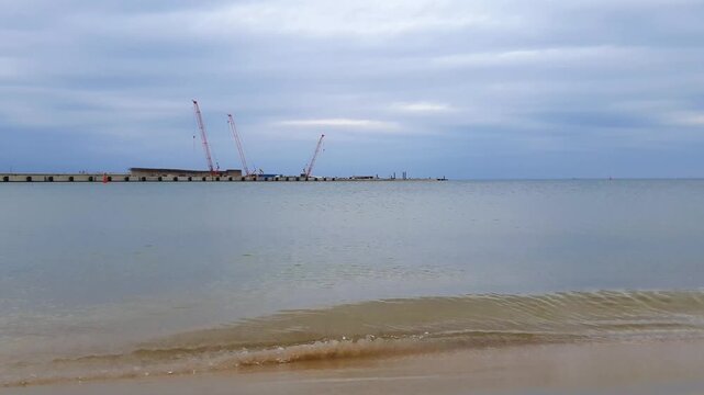 Empty seaport harbor on Baltic sea coast with calm coastal surf water waves rolling on sand beach and port quay on background under cloudy sky at overcast day. No people tranquil seaside landscape.