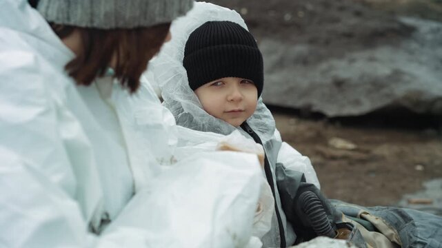 White child holding bread and blanket, child cradles sandwich, looks toward woman caregiver in white suit, knitted hat, muddy shore backdrop, intimate moment of shelter and comfort, quiet resilience