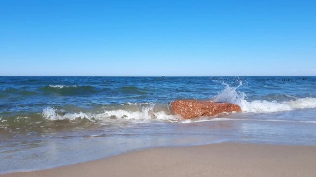 Tranquil seaside landscape of Baltic sea shore with calm turquoise coastal surf water waves rolling on boulder and empty sandy beach under clear blue sky at sunny day. No people seascape background.
