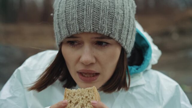 White female scientist eating ration outdoors near frozen shore, torn protective suit, beanie and muddy sleeve, biting stale bread with numb fingers, exhausted gaze, abandoned research site in gray