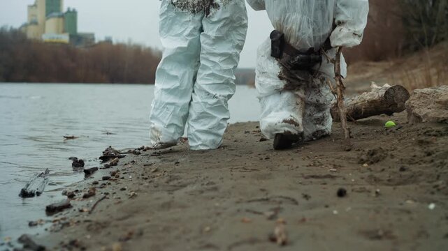 White technician and child inspecting shoreline, wearing protective suits and gloves while collecting water and soil samples, careful steps on muddy bank, detector and probe equipment in use,