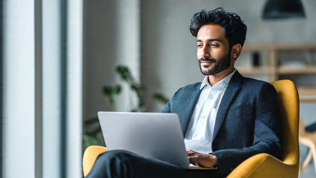 Indian male business executive sitting on a chair working on a laptop in a modern office, focused on remote studying, virtual training, or watching an online webinar.