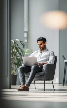 Indian male business executive sitting on a chair working on a laptop in a modern office, focused on remote studying, virtual training, or watching an online webinar.