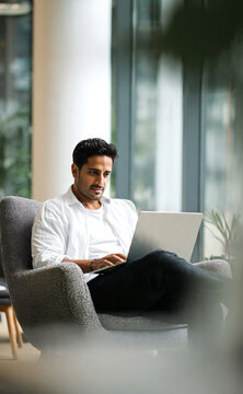 Indian male business executive sitting on a chair working on a laptop in a modern office, focused on remote studying, virtual training, or watching an online webinar.