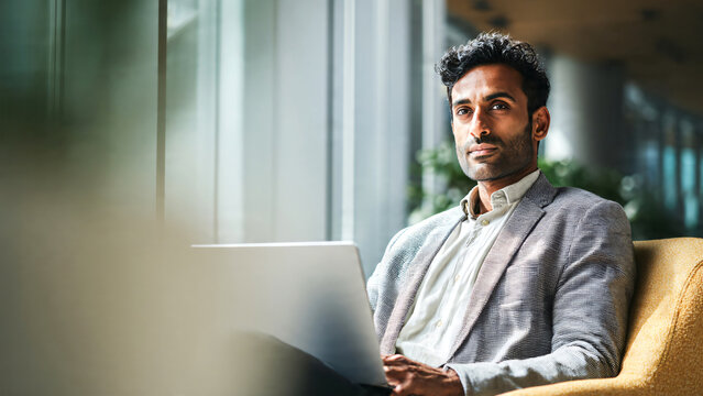 Indian male business executive sitting on a chair working on a laptop in a modern office, focused on remote studying, virtual training, or watching an online webinar.
