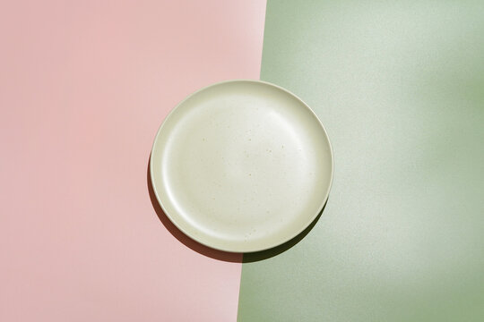 Minimalist flat lay of an empty ceramic plate on a geometric background split into pastel pink and sage green. Hard sunlight creates a strong shadow, giving a modern, graphic look. Top view