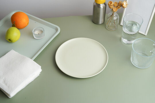 Minimalist table setting on a sage green background. An empty plate is ready for a meal, with fruit and water nearby. Clean, modern aesthetic with a calm mood. Perfect for wellness and food blogs