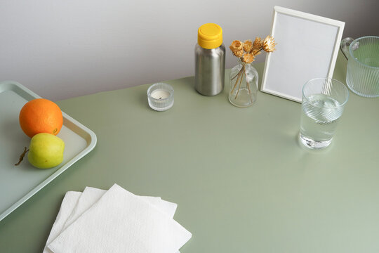 A modern and minimalist desk setup promoting a healthy lifestyle. This high angle view shows fruit, a reusable bottle, and a glass of water on a sage green table with a blank mockup frame