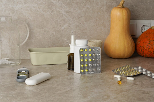 Still life of various medications, pills, vitamins, and a nasal spray on a beige kitchen counter. Autumn theme with pumpkins suggests seasonal health, colds, or flu. Muted colors, natural light