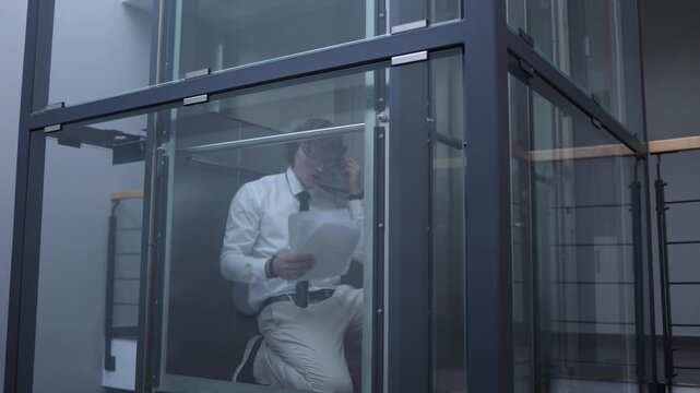 Office worker reviews documents and speaks through earphones inside a glass elevator capturing remote communication business focus multitasking and a modern flexible work routine