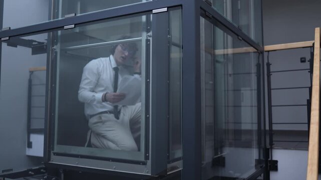 Office worker keeps reviewing documents and talking through earphones inside a glass elevator showing dedication overtime focus and the habit of working until the very end