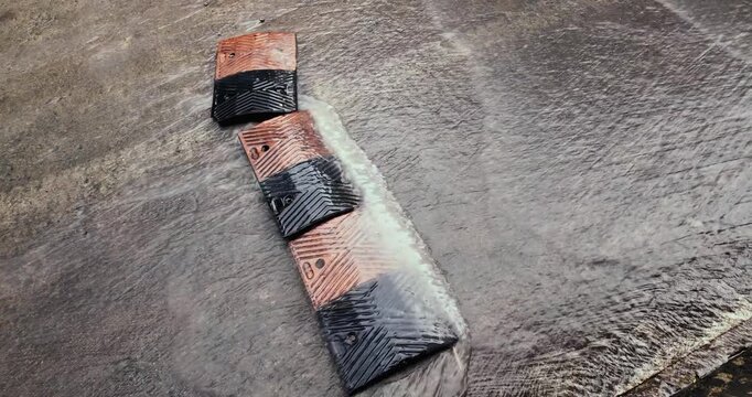 close up  of an orange and black rubber speed bump submerged in flowing rainwater. The footage shows street flooding and fluid dynamics as water rushes over the textured surface and chevron pattern 