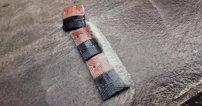slow motion of an orange and black rubber speed bump submerged in flowing rainwater. The footage shows street flooding and fluid dynamics as water rushes over the textured surface and chevron pattern 