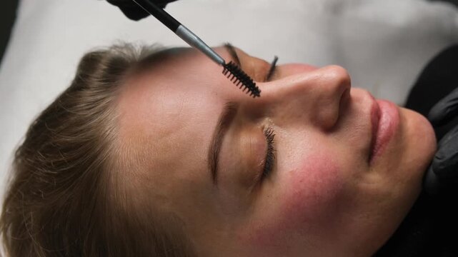 Professional beautician brushing a female client's eyebrows during a permanent makeup procedure