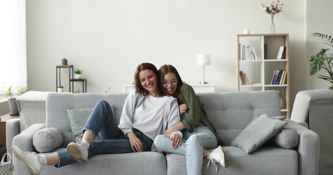 Cheerful mother and teen daughter jump on sofa and sit down, hugging, enjoying relaxation and conversation together in living room at home. Family bond, intergenerational connection, relationships