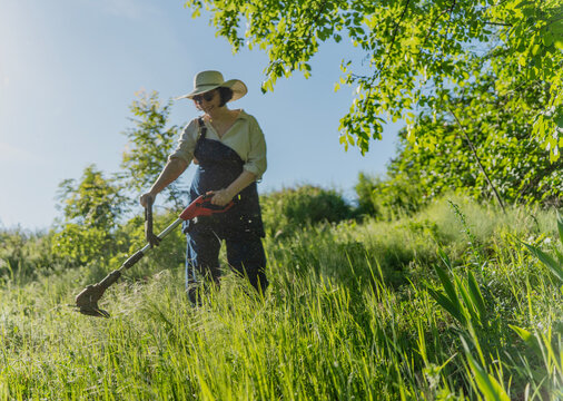 Mature woman mowing lawn with trimmer and flying grass in soft focus