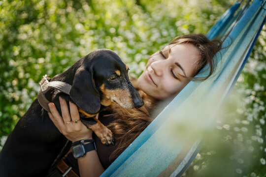 Happy young woman relaxing in hammock and holding cute dachshund dog in summer garden