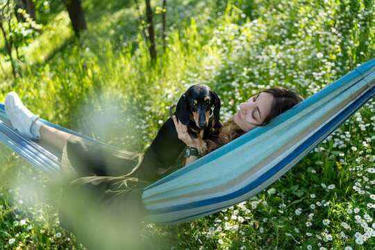 Happy young woman relaxing in hammock and holding cute dachshund dog in summer garden