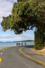Fototapeta premium A scenic coastal road curves towards a wooden pier extending into the calm blue water under a partly cloudy sky in Maraetai, Auckland, New Zealand.