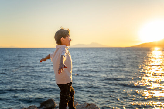 Happy caucasian boy standing on sea shore with arms outstretched at sunset enjoying summer freedom