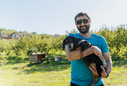 Happy mid adult caucasian man in sunglasses holding cute dachshund dog on summer field background
