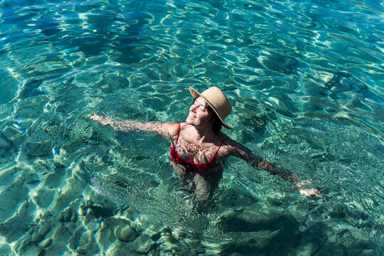 Top view of happy body positive brunette woman in red swimsuit and straw hat relaxing in turquoise sea