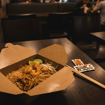 Shrimp noodle takeout box with lime wedge, bean sprouts, and crushed peanuts on a wooden table in a restaurant interior, emphasizing casual dining and ready-to-eat Asian food service