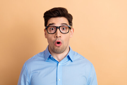 Surprised young man in glasses and blue shirt showing a shocked expression against a beige background