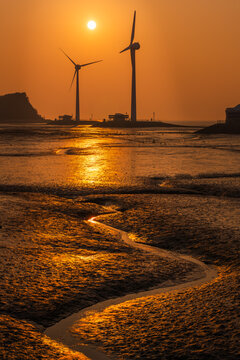 Dramatic sunset at Tando Port in Ansan, South Korea, featuring wind turbines and the Nue Island Lighthouse Observatory with golden light reflecting on the winding mudflat water trail.
