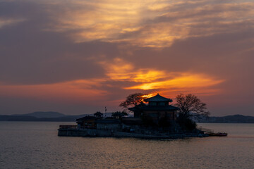Obraz premium Serene sunset view of Ganworam Hermitage, a unique Buddhist temple on a tiny island in Seosan, South Korea, with the glowing sun and vibrant orange sky reflected on the sea.