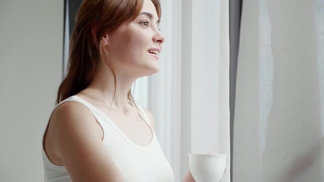 Happy caucasian woman open windows and drinking coffee in morning in bedroom at home, enjoy beverage with cozy for wellness, beautiful woman relax with beverage, self care, relaxation and lifestyle.