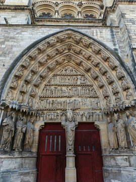 Frontal view of a monumental portal at Notre-Dame de Reims Cathedral, featuring classic red doors and a richly decorated tympanum. The shot highlights the vertical symmetry and the vast array of carve
