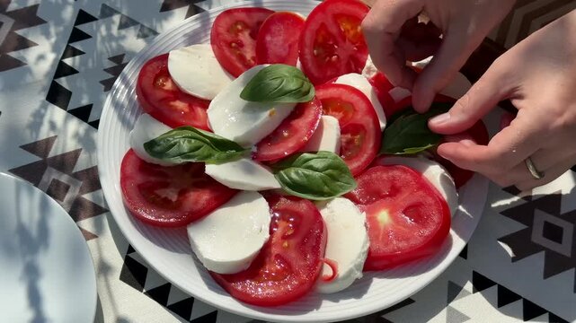Fresh caprese salad on a patterned table, sliced tomatoes and mozzarella with basil leaves, hands arranging the plate in bright natural light