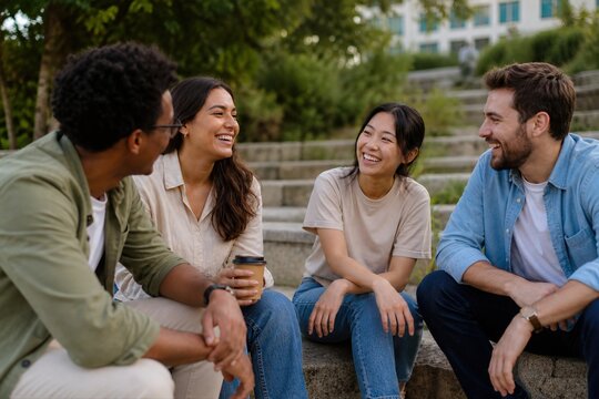 Group of diverse young multiethnic friends sitting on stone steps outdoors, talking and laughing together during a casual hangout in a city park on a sunny day.