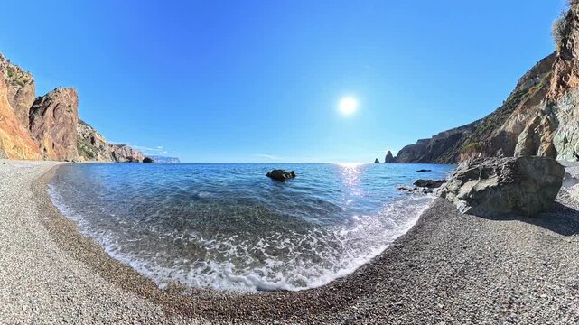 Beach cove ocean scene with crystal clear water and sunlight reflecting on a peaceful pebble shore with scenic mountain cliffs