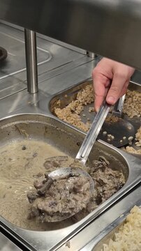 Cafeteria serving food worker scoops beef stroganoff with creamy mushroom sauce into a plastic container at a busy buffet lunch line