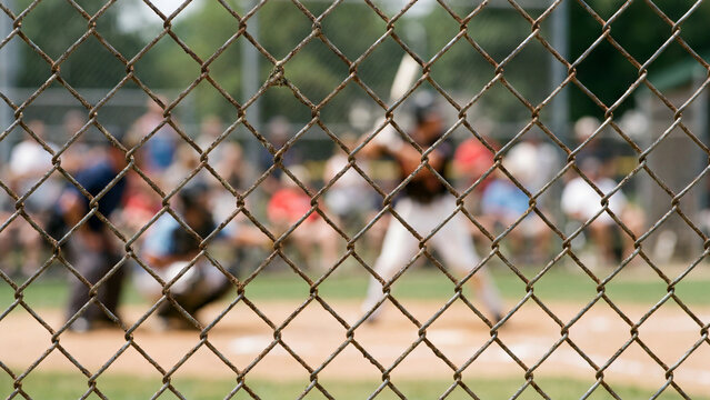 Baseball game viewed through chain link fence with batter catcher and spectators on outdoor field during competitive match for sports recreation and community athletics