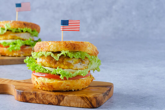 A festive bagel burger styled for Independence Day in the USA. Two bagel sandwiches on a cutting board, filled with crispy chicken, lettuce, and tomato on a gray background. space for text
