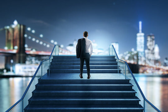 Businessman standing on wide stairs at night cityscape background facing bright light in modern creative scene symbolizing ambition and growth.