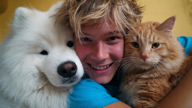 Young man with messy blond hair in blue t-shirt embracing a fluffy white Samoyed dog and an orange tabby cat, warm yellow background, close-up