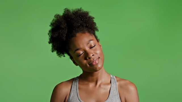 A young woman with dark skin and curly hair playfully sticks out her tongue and closes her eyes in a green studio for a video recording.
