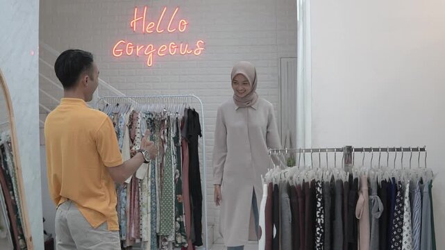 Woman in headscarf tries tunic in cozy boutique under neon hello gorgeous sign, male companion watches while stylist offers fit