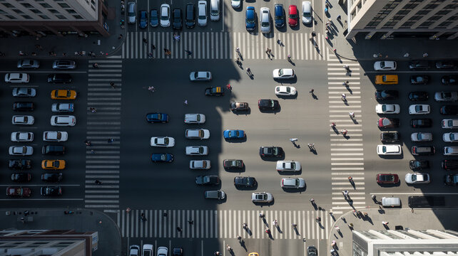 Aerial view looking down at busy urban street intersection featuring diverse cars driving and pedestrians walking across marked crosswalks in metropolitan city during bright sunlit afternoon hours.
