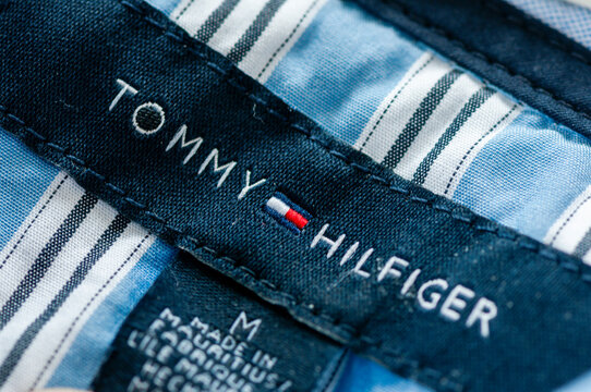 Close up macro shot of a navy blue Tommy Hilfiger clothing label stitched onto a light blue striped men's cotton shirt displaying the embroidered brand name, logo flag, and size tag.