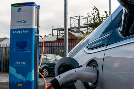 Title: A silver Vauxhall Ampera plug-in hybrid vehicle parked at an outdoor public charging station, connected to an electric cable for recharging in a UK urban parking area.