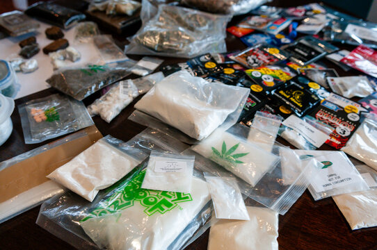 A diverse collection of illegal narcotics and synthetic legal highs, featuring white cocaine powder, brown heroin, and dried marijuana alongside branded foil packets of psychoactive research chemicals