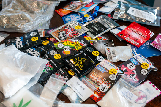 A diverse collection of illegal narcotics and synthetic legal highs, featuring white cocaine powder, brown heroin, and dried marijuana alongside branded foil packets of psychoactive research chemicals