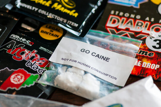 A diverse collection of illegal narcotics and synthetic legal highs, featuring white cocaine powder, brown heroin, and dried marijuana alongside branded foil packets of psychoactive research chemicals